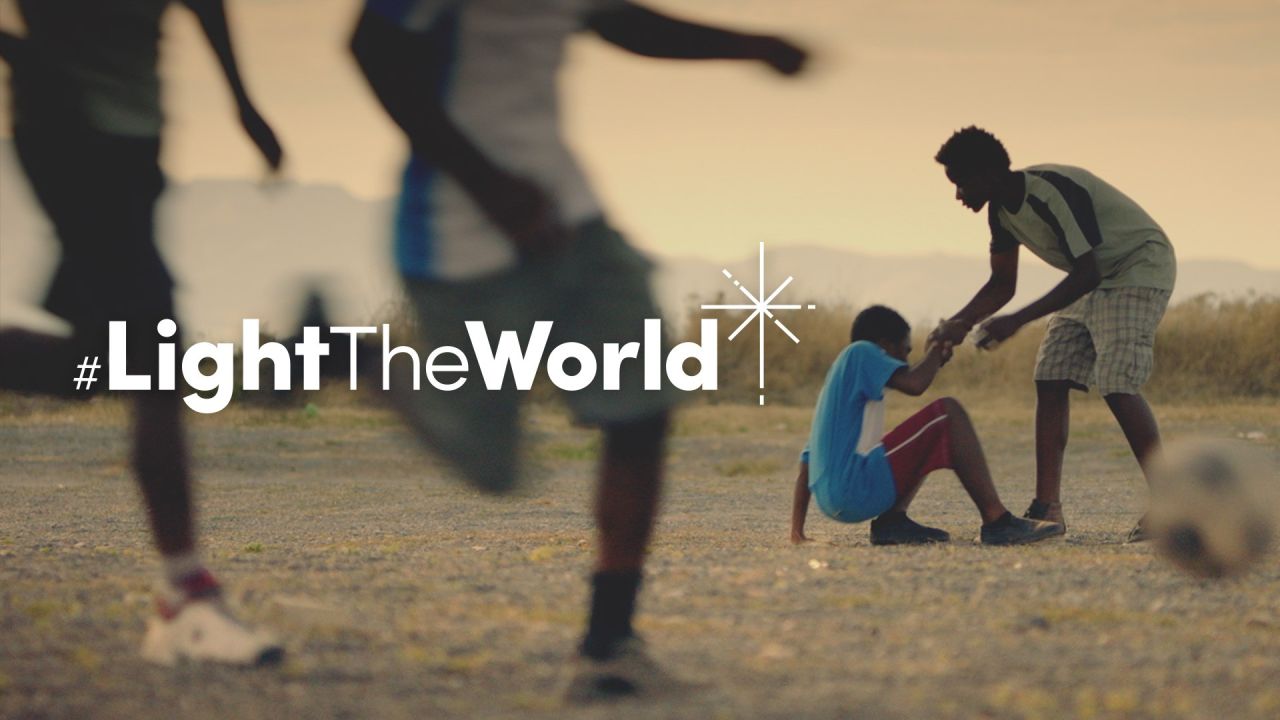 Poika auttaa toista poikaa nousemaan ylös tämän kaaduttua jalkapalloa pelatessaan Valoa maailmaan -videossa